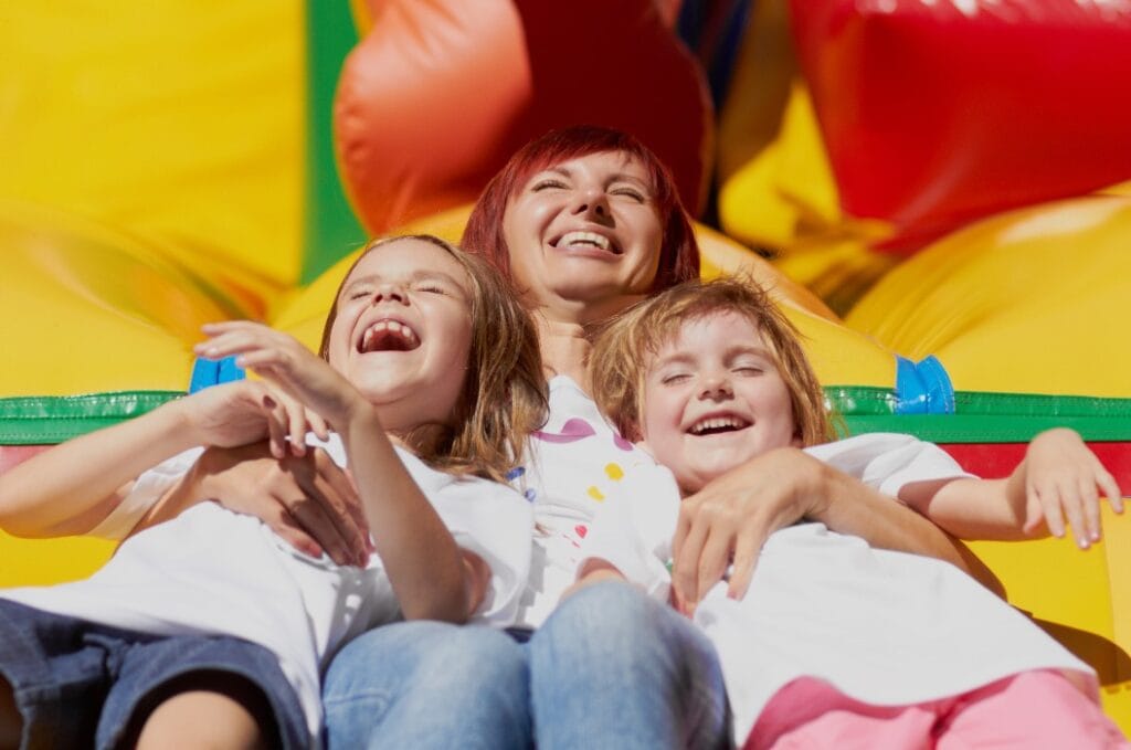Mother & her daughters having fun on jumping castle.Happy young adult mom with two kids laughing out loud with toothy smile.White childern enjoy summer with belove mother.Happy family portrait outdoor