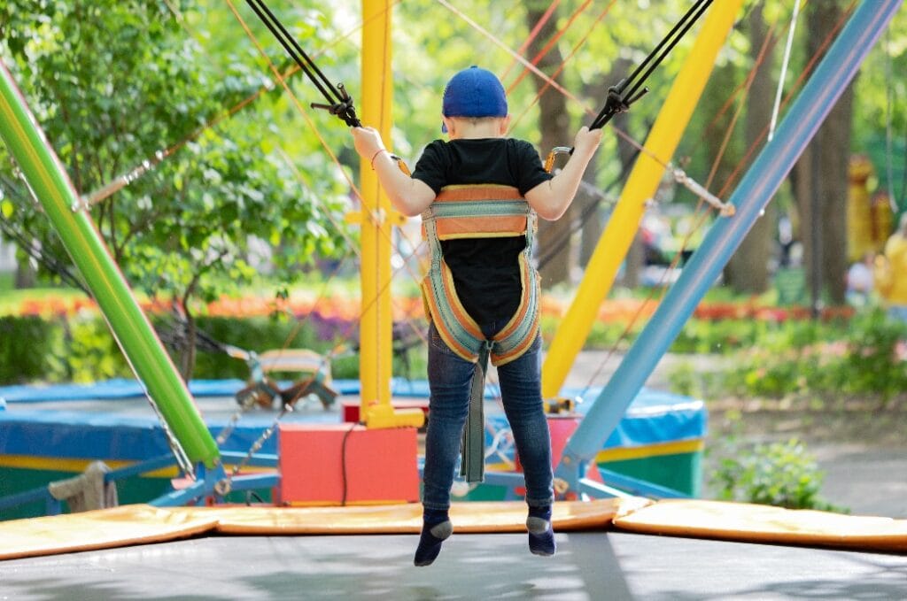 European boy is jumping on bungee trampoline in a Luna park. Back view.