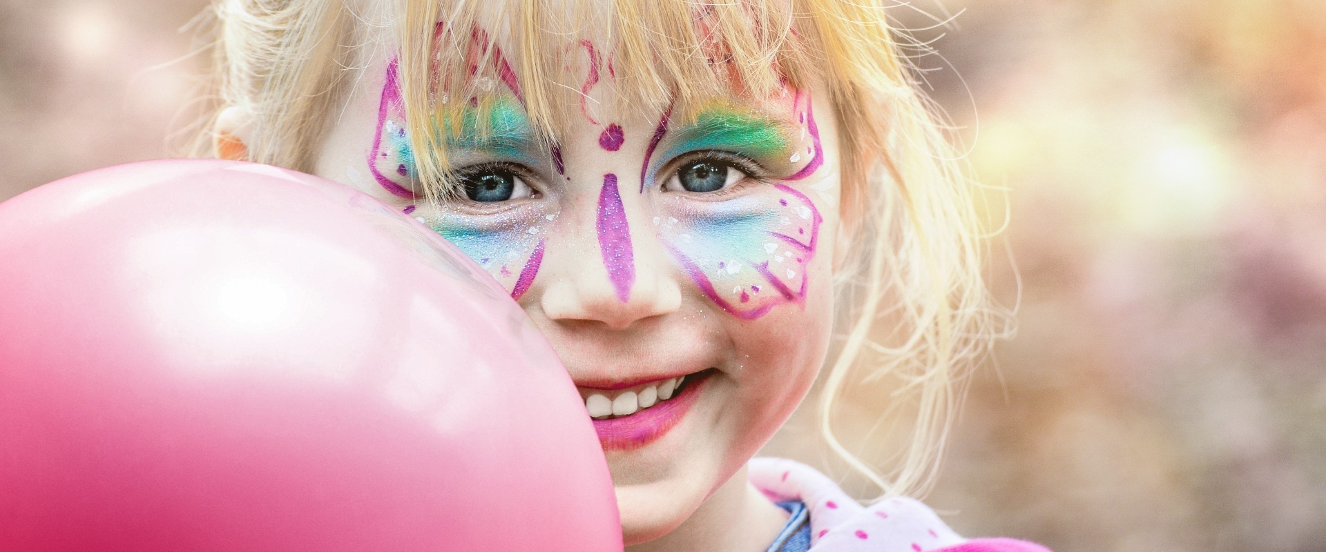 Glückliches geschminktes Mädchen beim Kinderkarneval auf einem Freizeitpark mit einem pinken Luftballon