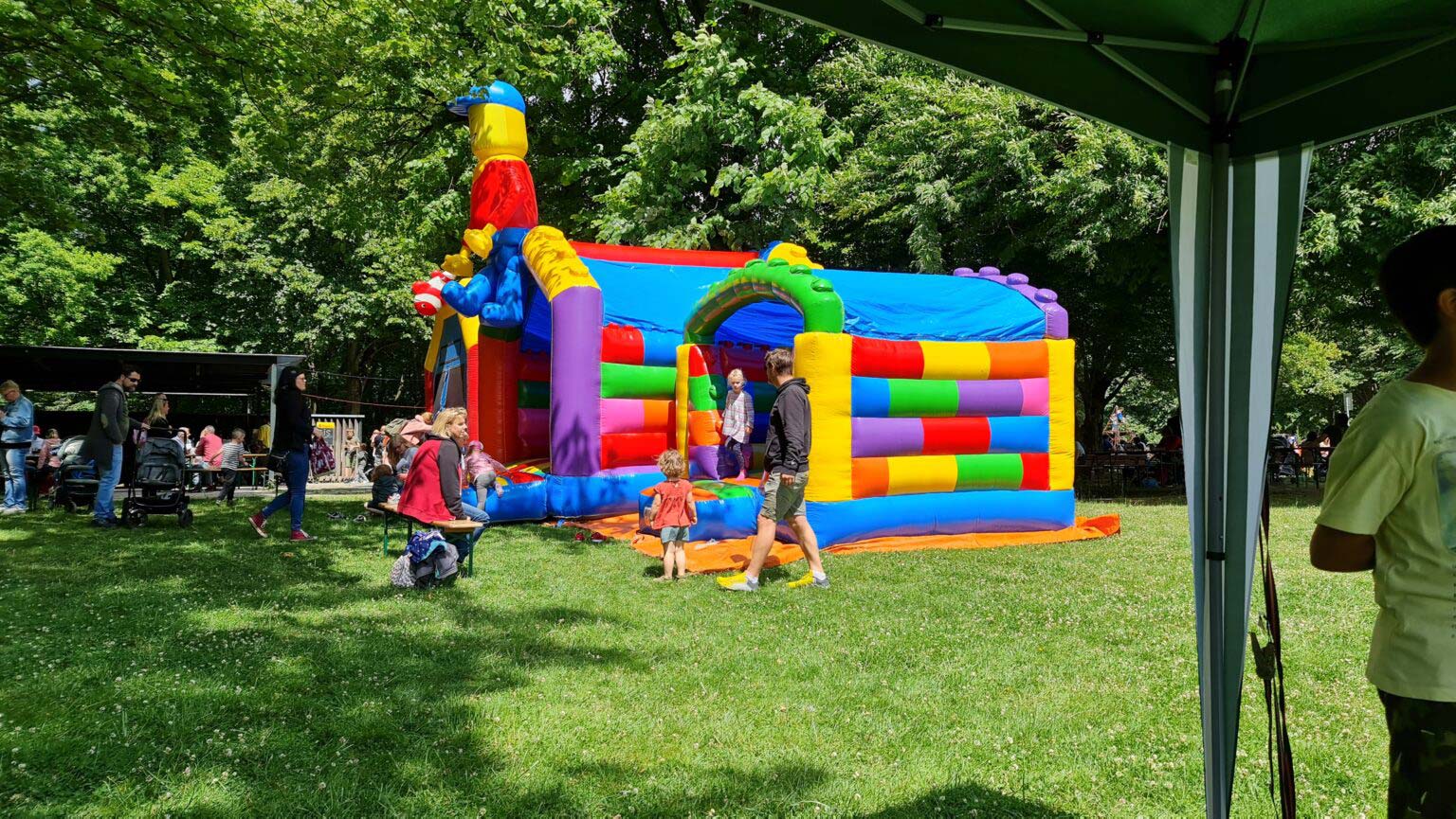 Eine bunte Hüpfburg im Freien, umgeben von spielenden Kindern und Erwachsenen im Park.