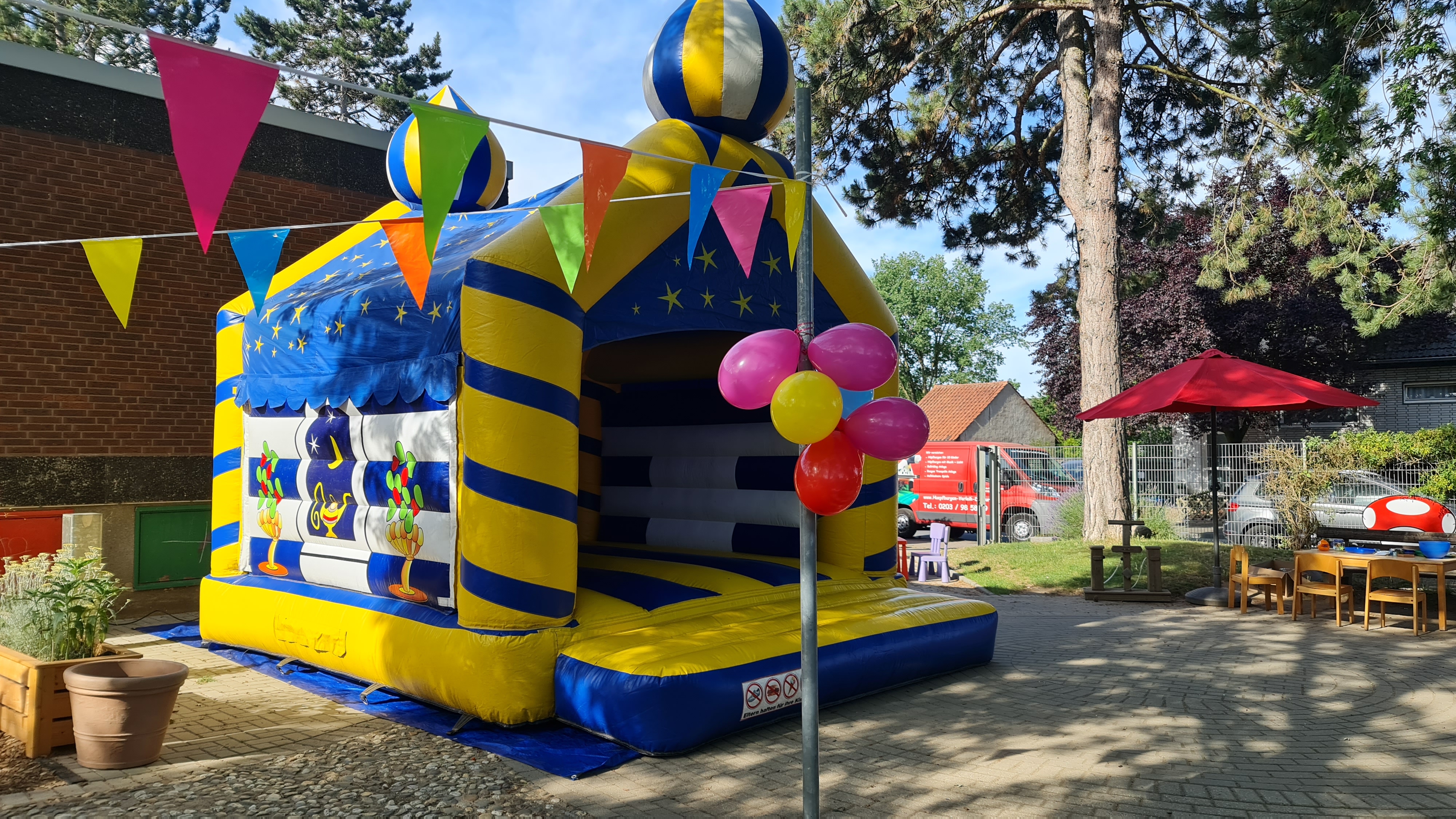 Eine bunte Hüpfburg im Freien mit blauen und gelben Streifen. Luftballons und Girlanden schmücken sie.