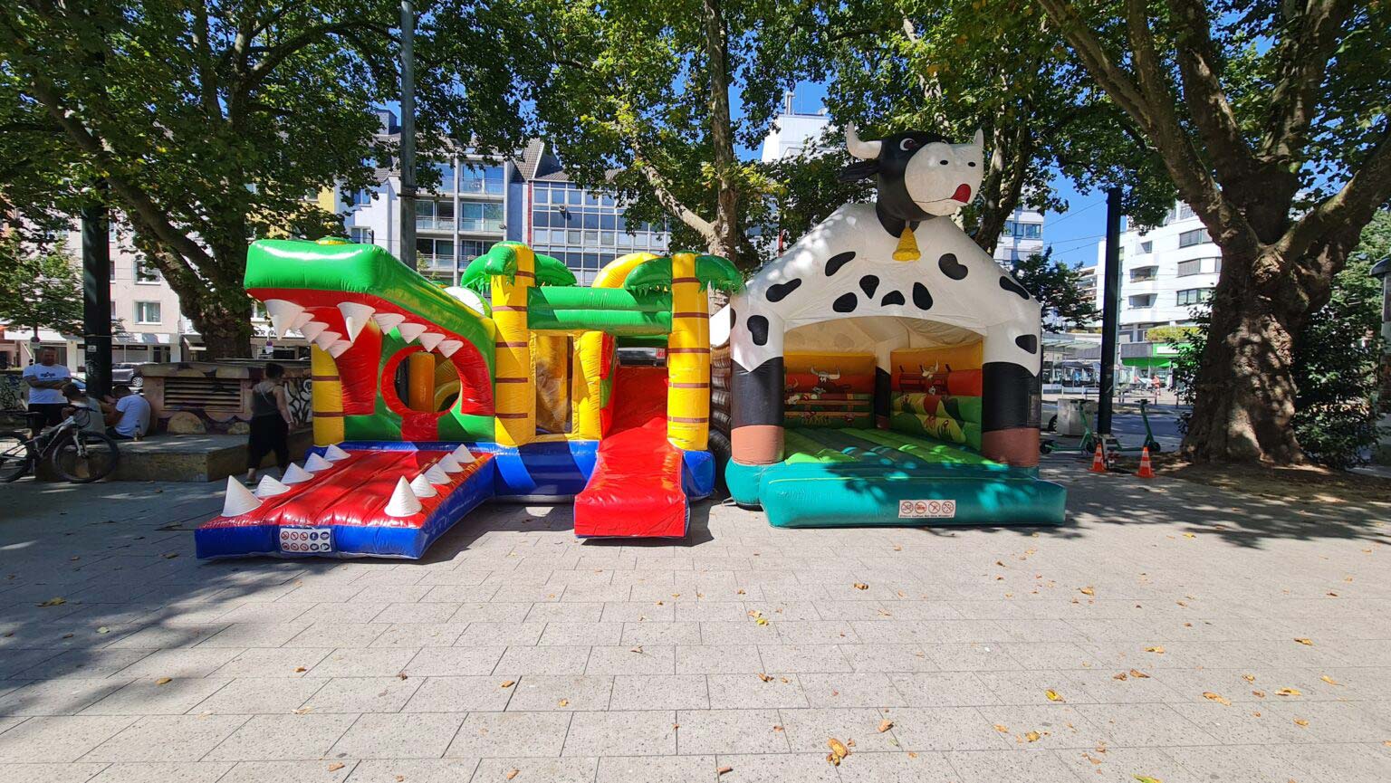 Drei bunte Hüpfburgen: Krokodil, Kuh und Burg, auf Platz unter grünen Bäumen; sonniger Tag.