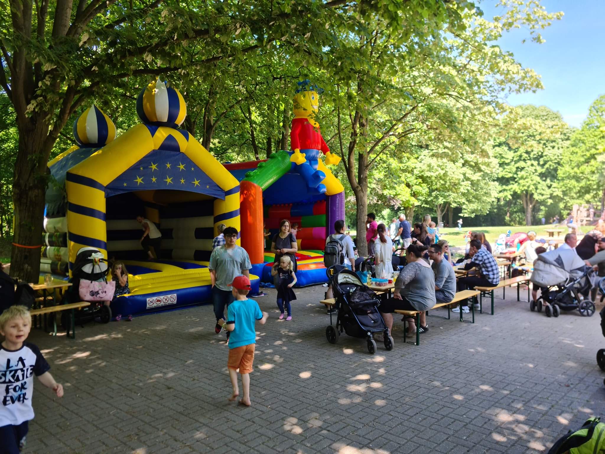 Ein Park mit Hüpfburgen, Kindern beim Spielen, Erwachsenen auf Bänken, viele Bäume, sonnig.
