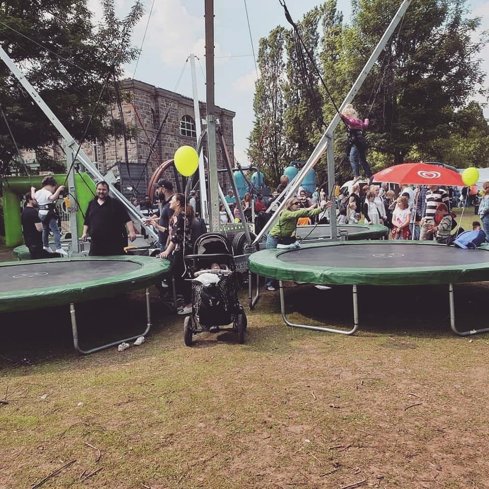 Menschen genießen eine Messe mit Trampolinen, Sonnenschirmen und bunten Luftballons im Freien.