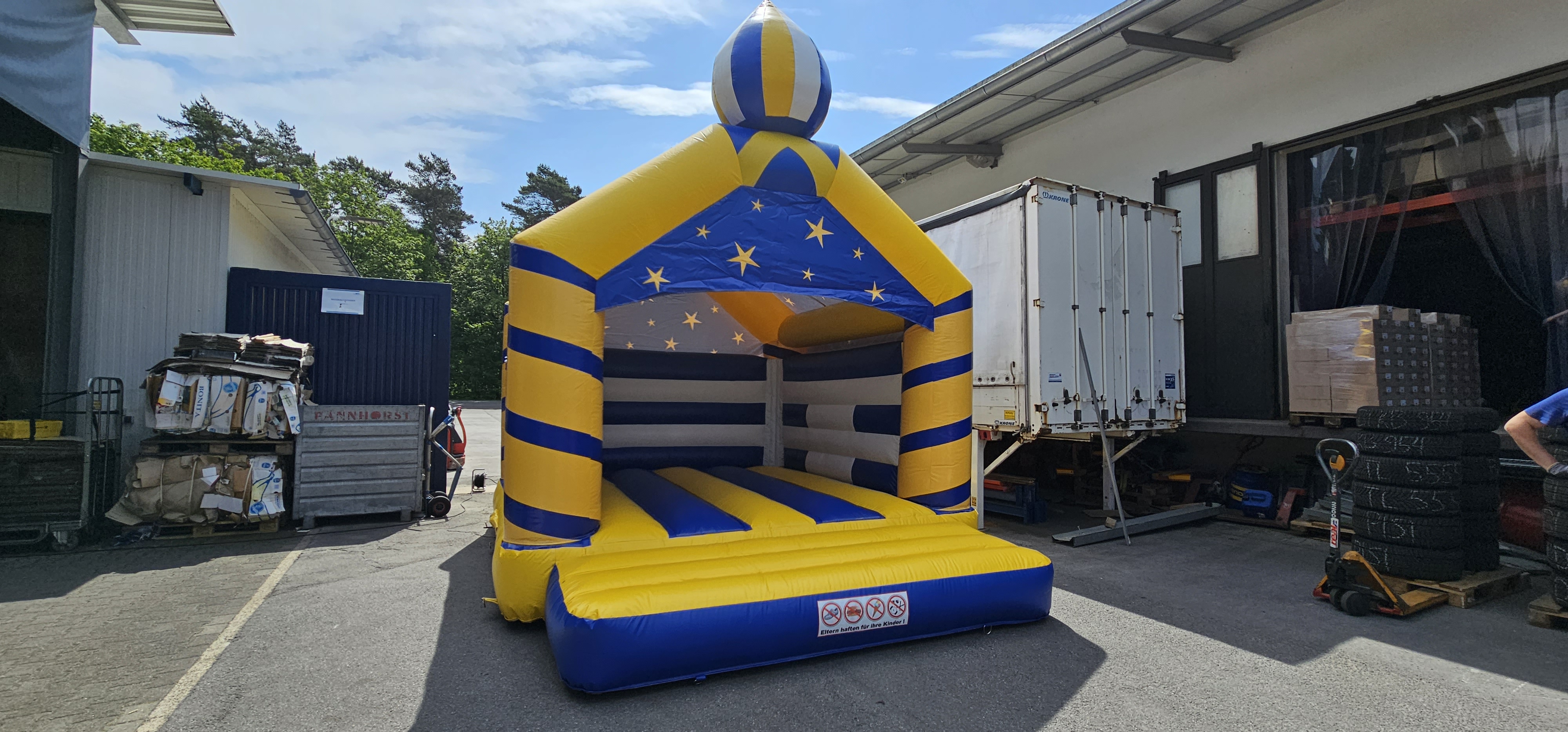 Gelbe und blaue Hüpfburg mit Sternenmuster, umgeben von Lagerbereich und Bäumen im Hintergrund.