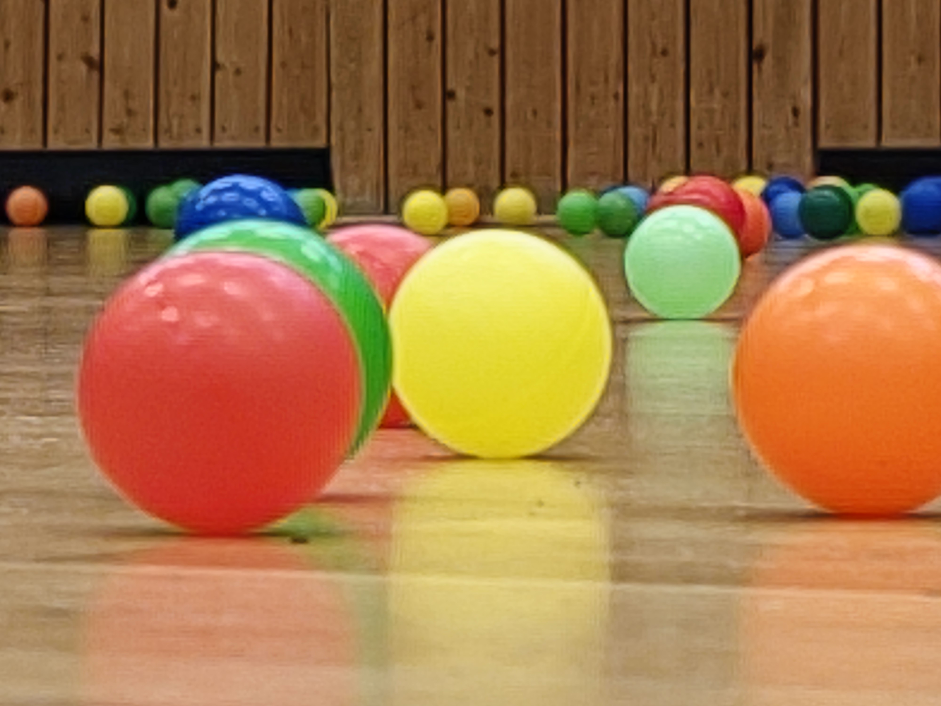 Bunte Plastikbälle liegen auf einem Holzboden in einer Sporthalle, teils unscharf im Hintergrund.