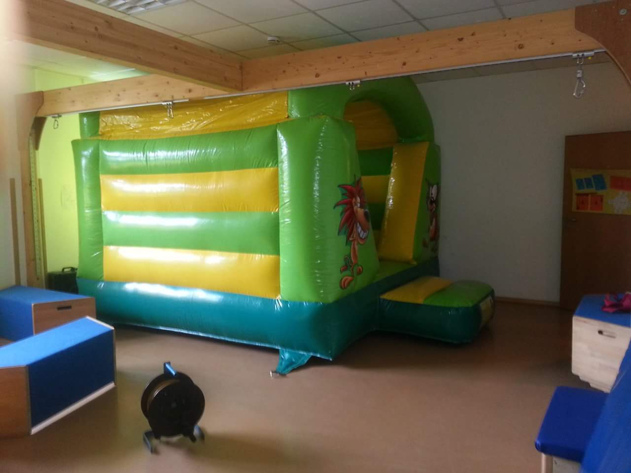Aufblasbare grüne und gelbe Hüpfburg mit Cartoonfiguren, steht in einem Raum.