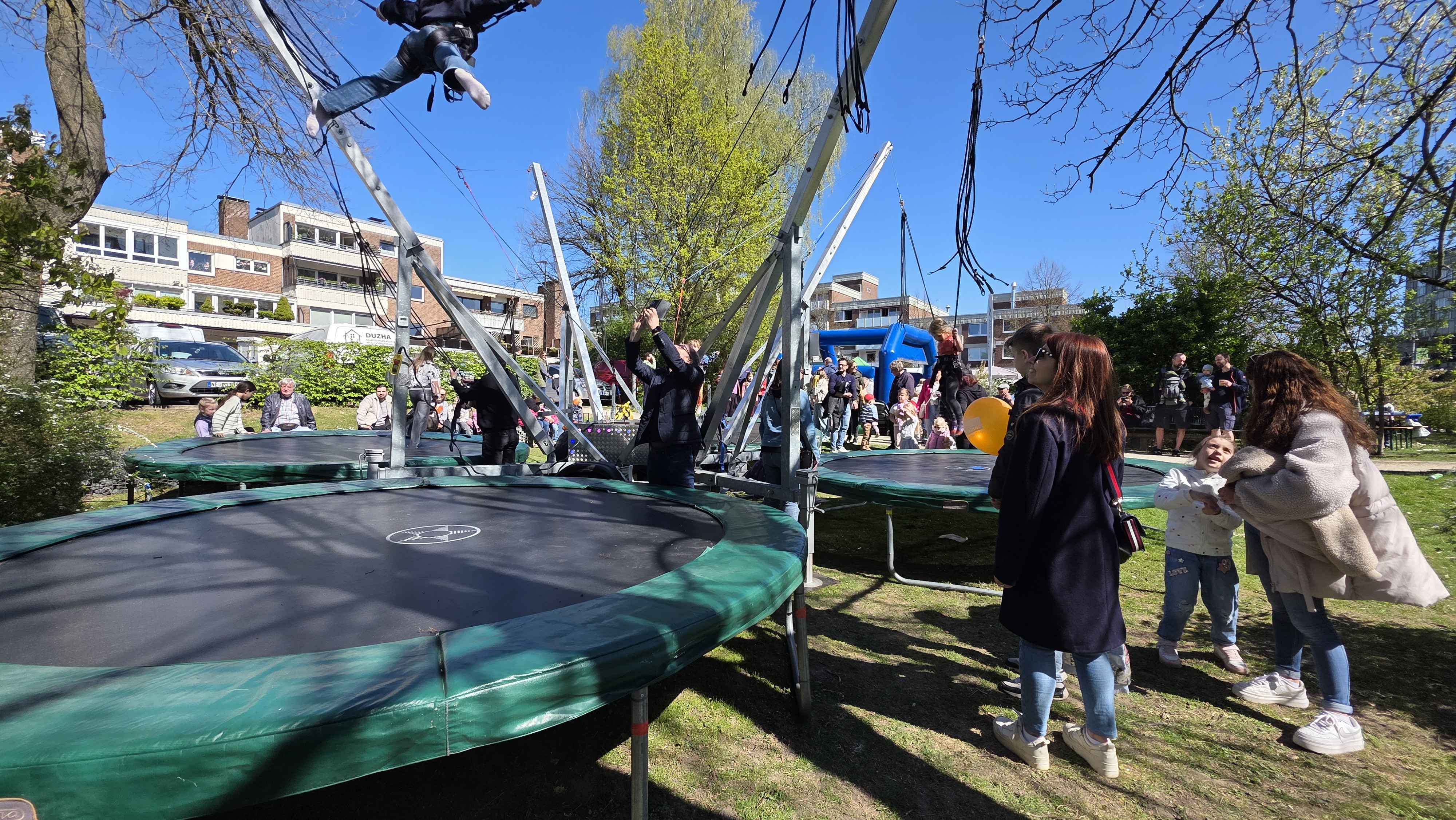 Eine Parkszene mit Menschen auf Trampolinen bei sonnigem Wetter; lebhafte und fröhliche Stimmung.